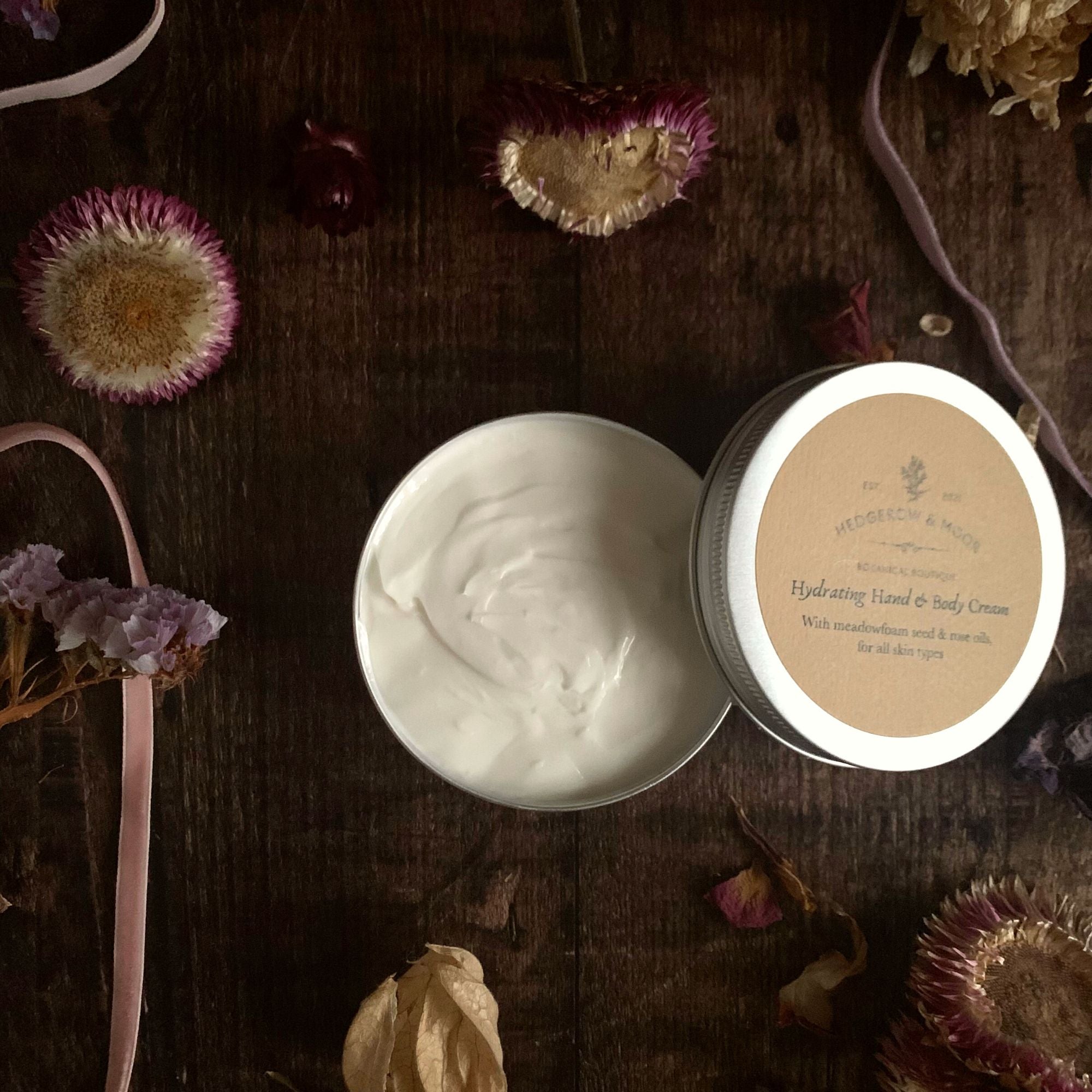Open jar of Hedgerow and Moot `body cream with lid on a wooden surface surrounded by flowers