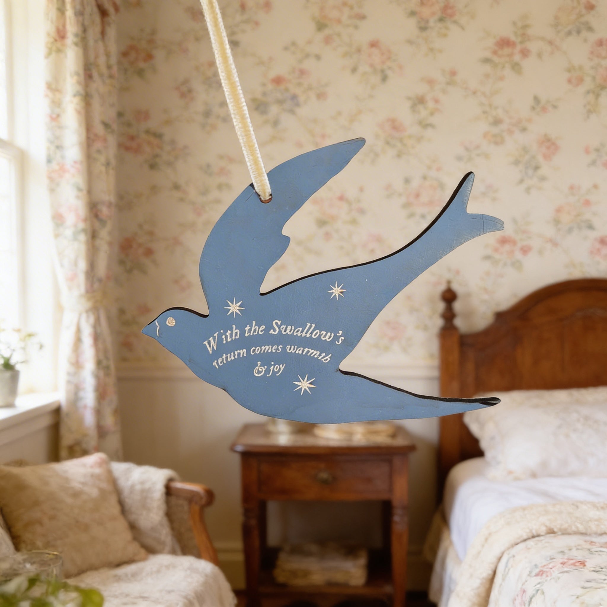 Decorative swallow decoration with text hanging in a room with floral wallpaper and wooden furniture.