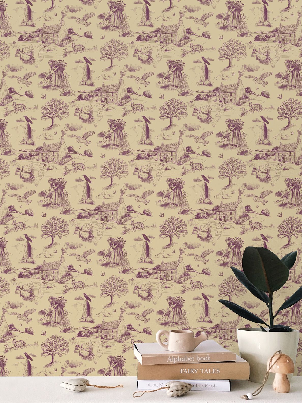 A styled interior scene showing a cream hedgerow and moor design wallpaper with a repeating purple toile-style print of animals, trees, and small houses. In the foreground, a white tabletop holds stacked books, a small ceramic mug, a potted plant, and a wooden mushroom-shaped decor object.