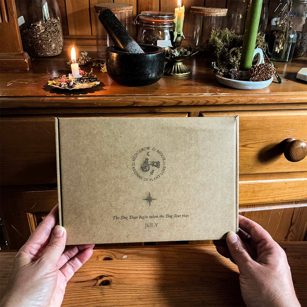 Person holding a hedgerow and moor monthly subscription  box with a circular design on a wooden surface with candles and jars.