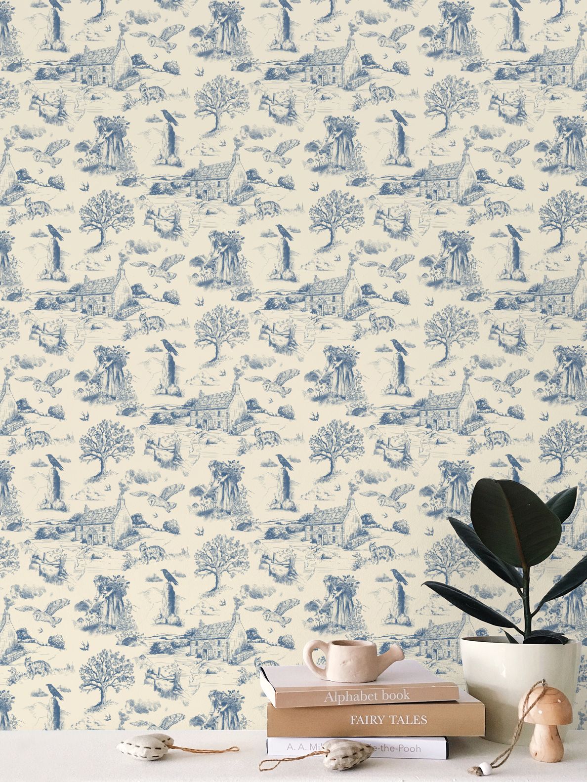 Blue and cream toile wallpaper with a repeating whimsical pattern of animals, trees, and houses, styled on a wall above a tabletop with books, a plant, and small decorative objects.