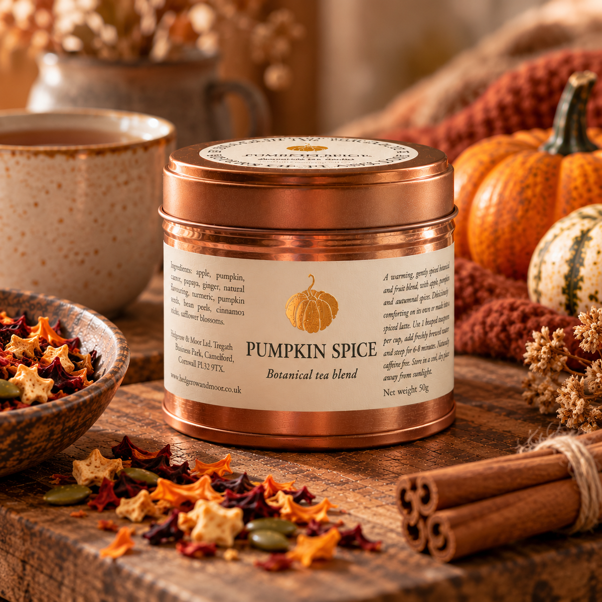 A copper-toned metal tea tin labeled 'Pumpkin Spice Botanical tea blend' sits on a rustic wooden surface surrounded by autumn props including dried leaves, cinnamon sticks, a mug, and pumpkins.