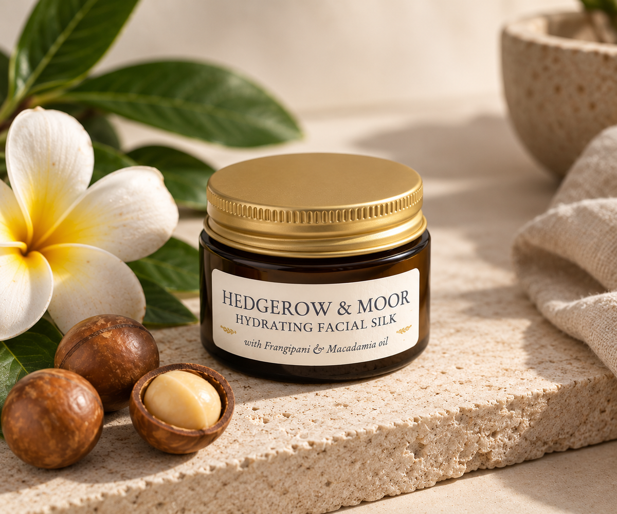A small amber glass jar of Hedgerow & Moor Hydrating Facial Silk with a gold lid and white label, styled on a stone surface with a white flower, green leaves, and macadamia nuts.