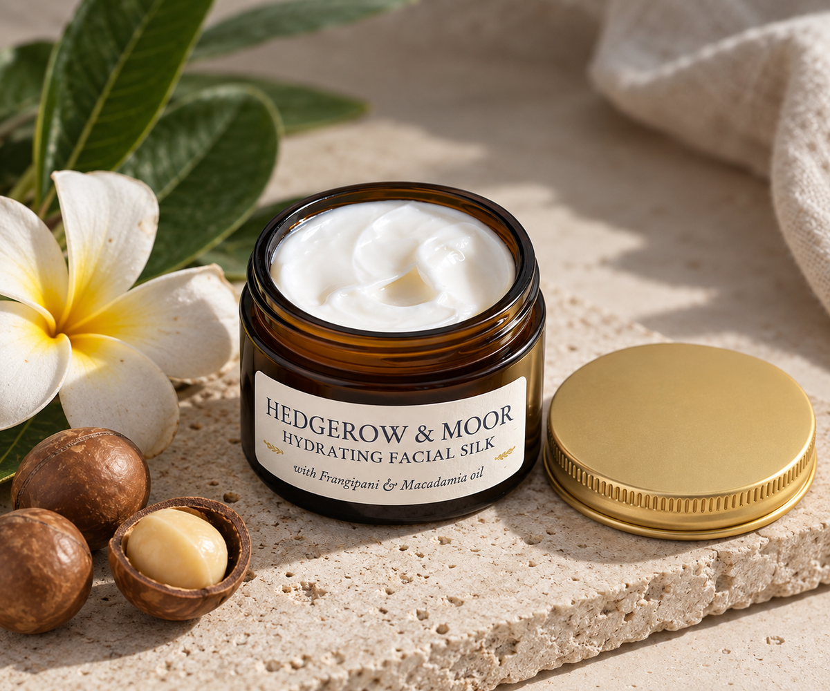 An open amber glass jar of Hedgerow & Moor Hydrating Facial Silk with visible white cream, shown on a stone surface with a gold lid beside it, plus a white flower, green leaves, and macadamia nuts as styled props.