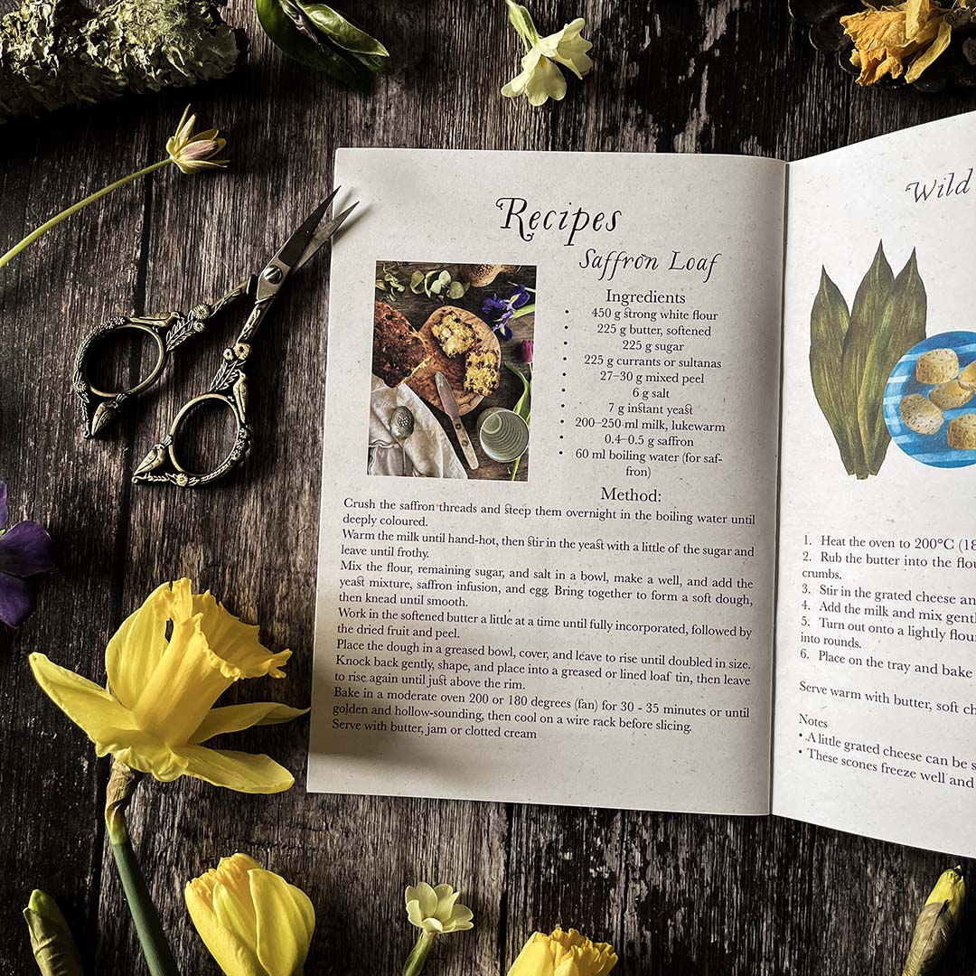 Open seasonal cookbook with saffron  recipe on a wooden surface with flowers and scissors.