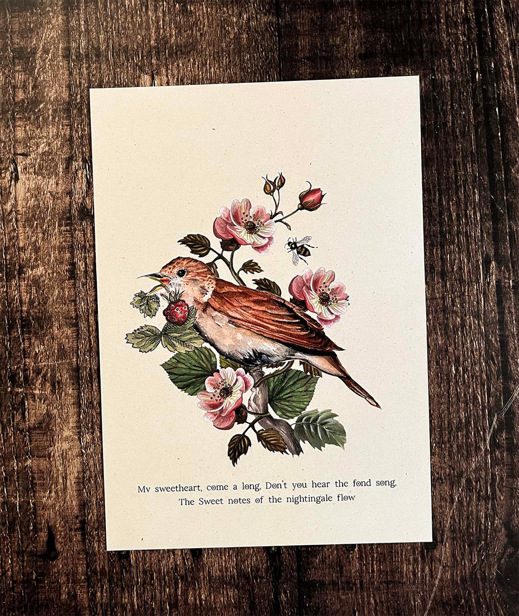 A vintage-style art print featuring a brown bird perched on a flowering branch with pink blossoms and green leaves, placed on a dark rustic wooden surface. The print includes printed text at the bottom.