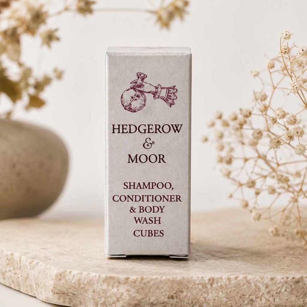 A tall rectangular box of Hedgerow & Moor shampoo, conditioner, body wash, and body wash cubes stands centered on a light stone surface with dried floral decor in the background.