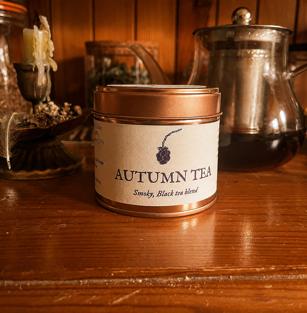 A tin of Autumn Tea, a smoky black tea blend, displayed on a rustic wooden table with a vintage teapot and candle in the background.