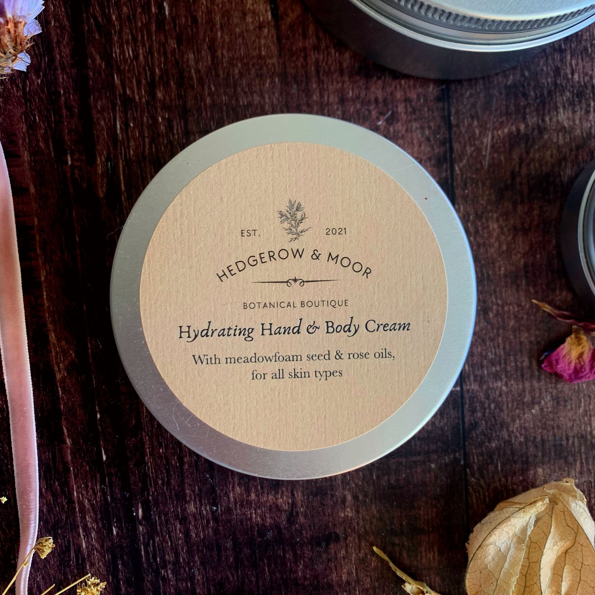 Hedgerow & Moor hydrating hand and body cream container on a wooden surface with dried flowers.