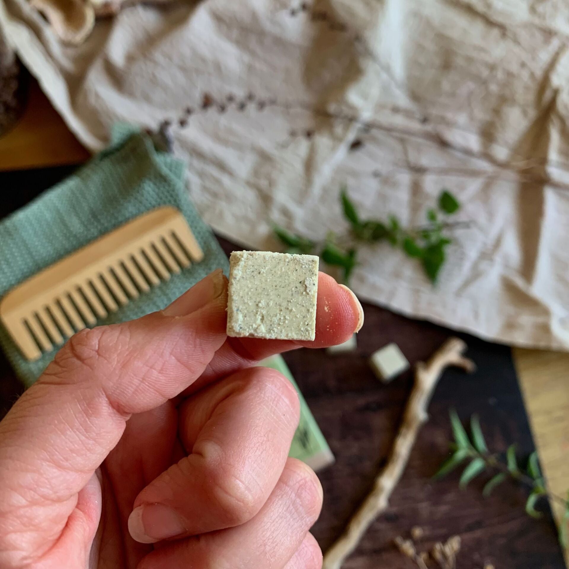 Hand holding a small shampoo cube for oily hair with a background of a textured fabric and a comb.