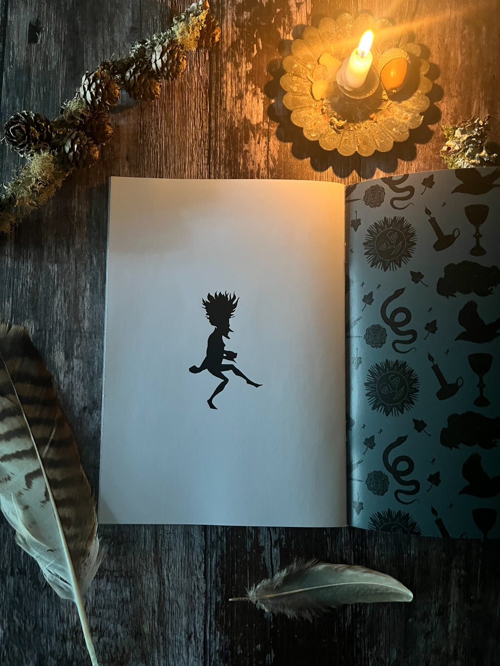 Open folklore zine showing a black spriggan silhouette and patterned inside cover, photographed by candlelight.