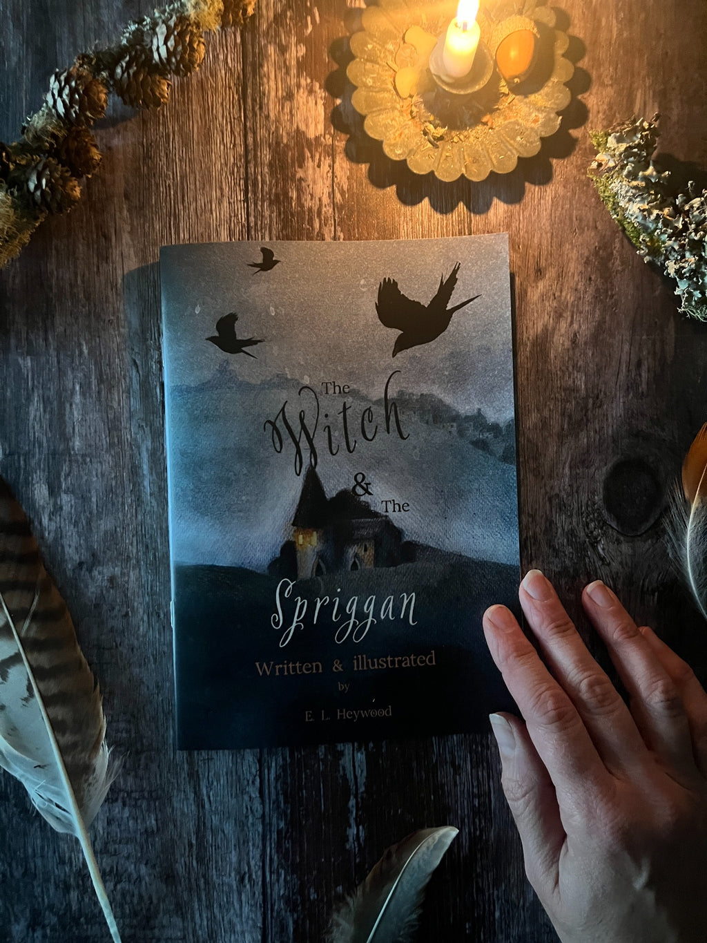 The Witch & The Spriggan booklet beside a lit candle, with a hand resting on its illustrated cover.